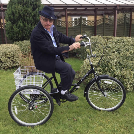 A gentleman wearing a dark jacket, hat, and glasses sits on a black electric tricycle with a white rear basket.