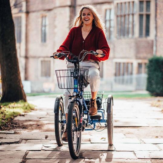 A smiling woman with long blonde hair, wearing a red sweater and white pants, rides a blue electric tricycle with a front basket along a sunny stone path near a historic building.